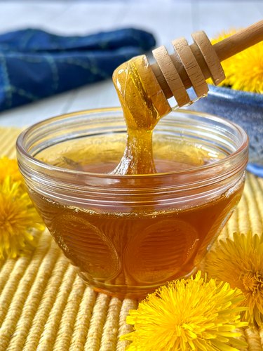 Homemade dandelion honey in a glass jar with a blue pottery bowl full of fresh dandelions