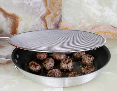 splatter screen over meatballs in a frying pan on a stone countertop