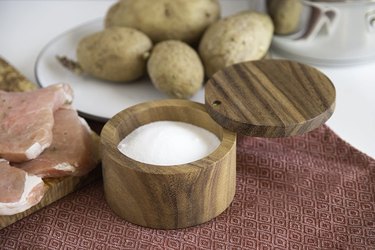 Dark wood round salt cellar with a lid that swings open or closed on a countertop next to uncooked meat and potatoes.