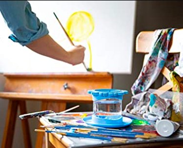 A person painting at an art easel with a paint brush cleaner rinse cup.