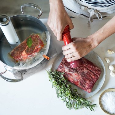 Breville immersion circulator cooking a steak in a stainless pot, while a chef seasons a second piece of meat
