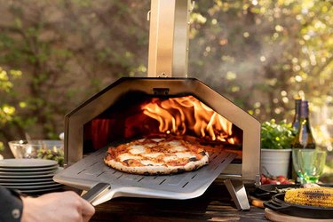 Ooni peel pulling a baked pie from an Ooni pizza oven in a back yard, with dishes on either side and trees in the background