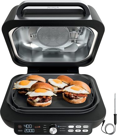 Ninja Foodi smart grill, shown on a white ground with the lid raised and egg-topped bacon cheeseburgers on the cooking surface