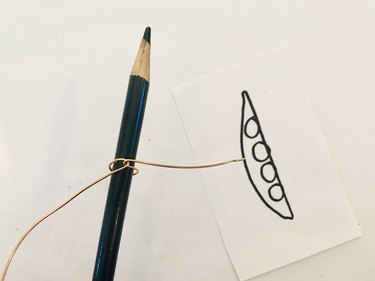 An image of twisted wire wrapped around a colored pencil; the tip of the pencil is pointing up. There is a drawing of a pea pod in the background