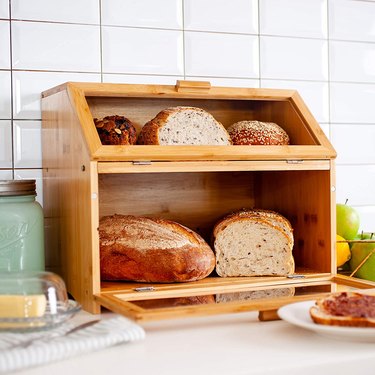 Two-tier bamboo bread box on a counter containing breads, bagels, muffins, and more.
