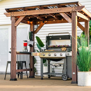 Pergola-style grill gazebo with cedar frame and steel roof, shown covering a grill on a deck with decorative plants alongside