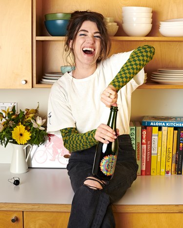 Caucasian woman sitting on kitchen counter opening a bottle of champagne