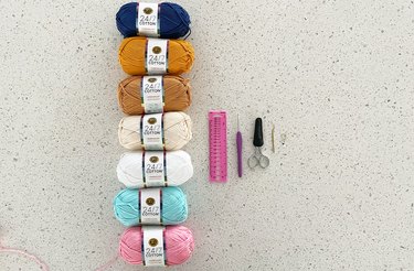 Seven skeins of colorful yarn, a small pink plastic ruler, a purple crochet hook, a pair of embroidery scissors, a gold darning needle and a stitch marker