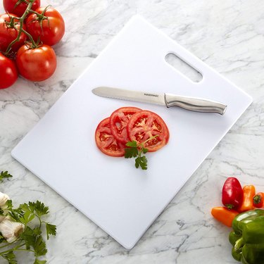 Farberware Plastic Cutting Board on a Marble Countertop Being Used to Slice a Tomato With a Serrated Knife
