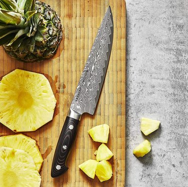 The Kramer by Zwilling Euroline Damascus Collection 8-in. Narrow Chef's Knife on a wood cutting board surrounded by chopped pineapple.