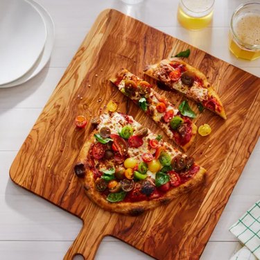 Olive wood cutting board, shown with a cut-up, colorful flatbread pizza on a pale tablecloth, with plates and beverages visible alongside