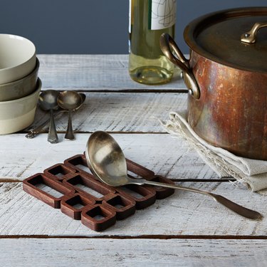 Mid-century modern inspired wood trivet on a wood slat table next to a copper pot, soup bowls, and a wine bottle.