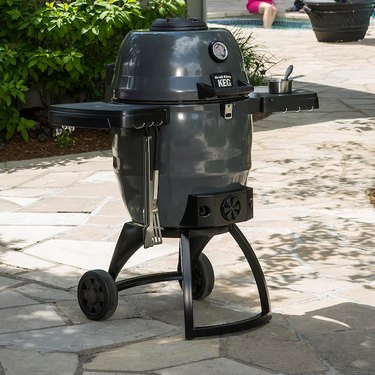 Broil King Keg 5000 kamado cooker, shown on a stone patio with shrubs and a swimming pool in the background