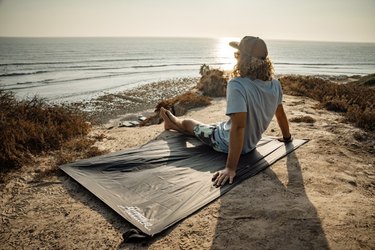 Man sitting on water-resistant picnic blanket with his back facing the camera. He's sitting on a sandy perch overlooking the ocean and the sun is setting.