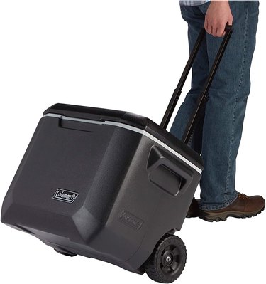 Coleman rolling cooler, shown with handle extended and a man pulling it, against a white ground