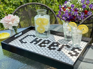 Black and white serving tray reading "Cheers" in tile, topped with two glasses of lemon water and a pitcher of lemon water