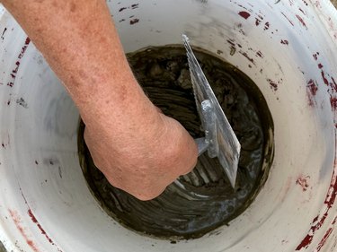Hand and metal tool mixing brown tile grout in a white bucket