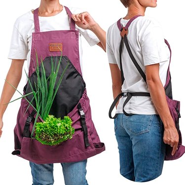 Anonymous woman wearing the Grace & August garden apron in the color "Aubergine." You can see the frontside and backside of the apron side-by-side. In the front, she is carrying lettuce and green onions.