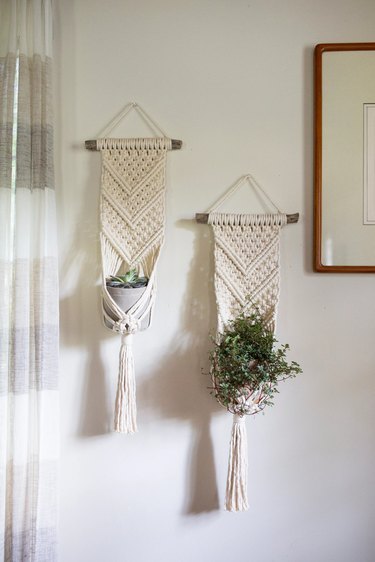 Two macrame plant hangers on a white wall. They're like traditional macrame wall hangings that transition into plant holders at the bottom. Each holder has a plant pot in it, although they're relatively small.
