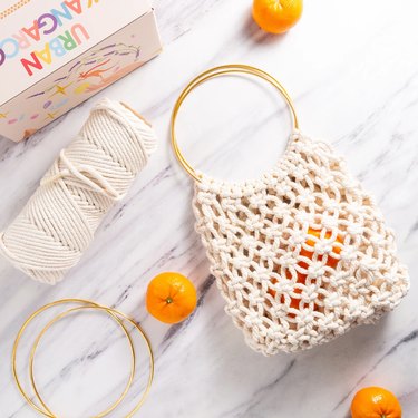 A macrame bag with gold hoop handles on a marble countertop. The bag is big enough to hold a couple of clementines. The photo also shows the materials that are included like the cotton rope and gold hoops.