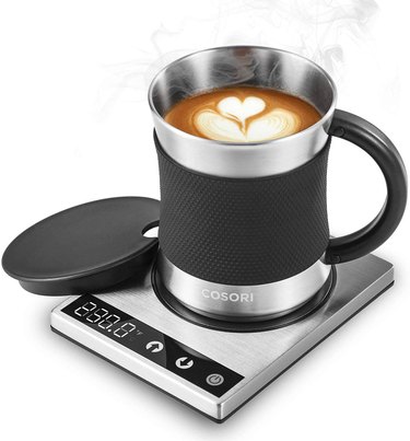 Stainless steel warming mug with black lid and a heat-protecting band around the middle sitting on a warming pad with a temperature display and controls.
