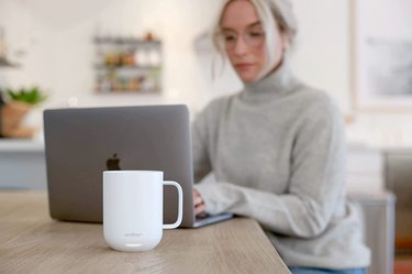Blonde woman at a desk working on a laptop next to a white warming mug by Ember.