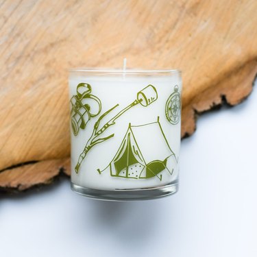 White candle in glass container featuring green decals in the shape of a stick with marshmallow, a camping tent, compass and binoculars