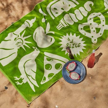 Green beach towel from Brooklinen with New York City motifs like pizza, a pigeon, a pretzel, and the Statue of Liberty in an off-white color.