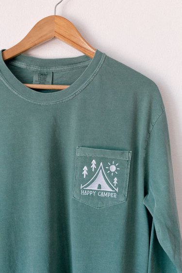 Green long-sleeved T-shirt with embroidered tent and trees alongside the words "Happy Camper" on pocket