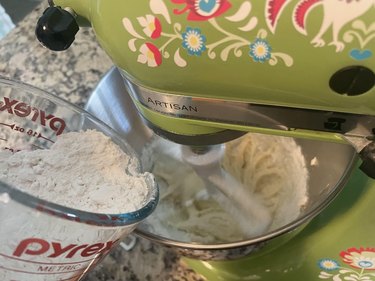 Flour in glass measuring cup being added to wet ingredients in Kitchen Aid mixer