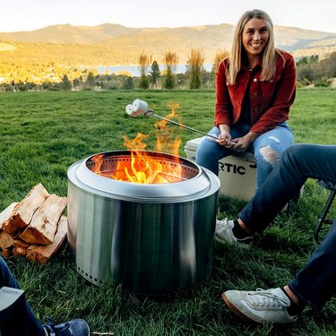Woman roasting a marshmallow over a Solo Stove in a grassy field next to a lake.
