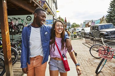 Couple walking down a city street past a bike shop. The woman is wearing a Hydro Flask water bottle sling across her body in the 'Snapper' colorway (a deep berry color).