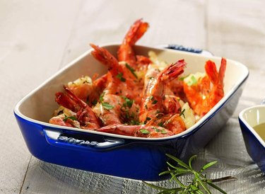 Blue Staub baking dish filled with shrimp, shown on a white tablecloth with a sprig of rosemary