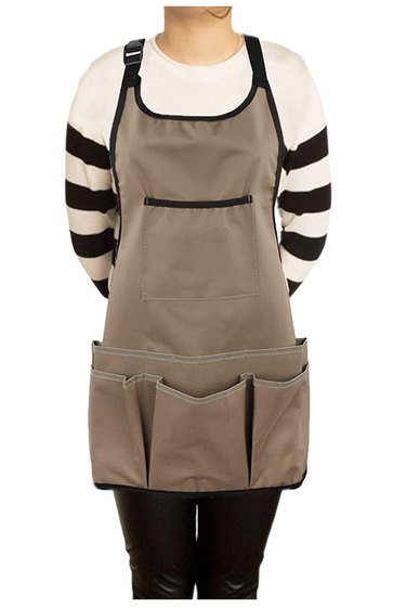 Anonymous woman wearing the PATILWON Gardening Apron with Pockets over a black and white striped sweater. The color is light brown and it has black trim and very large pockets.