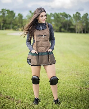 Young woman standing in a grassy field wearing the Typhon East garden apron and the included knee pads. The apron is a light brown color and has dark green accents on the three large bottom pockets.
