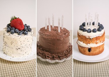 A poppy seed cake with lemon frosting topped with fruit, a chocolate cake topped with chocolate frosting and chocolate shavings, and a blueberry cake topped with vanilla frosting and blueberries