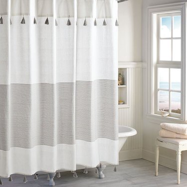Peri Home Panama Stripe Shower Curtain covering a clawfoot tub in a coastal-style bathroom. The shower curtain is tan, white and gray with very thin stripes and tassels near the top and bottom.