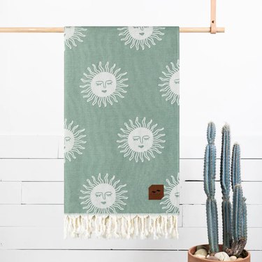 Slowtide Slow Burn Beach Blanket hanging from a wooden dowel and situated next to a cactus. The blanket is sage green with white rope fringe. The pattern is large and it's whimsical suns with faces (eyes closed).