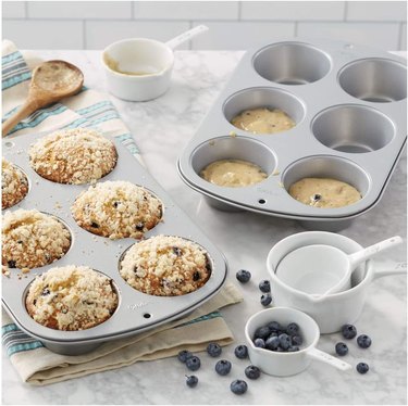 Two Wilton jumbo muffin tins on a marble counter, one with baked blueberry muffins and the other containing muffin batter; surrounded by measuring cups and blueberries