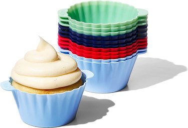 Stack of colorful OXO silicone baking cups in blue-green, dark blue, red and pastel blue, with a baked and iced muffin standing in front of the stack