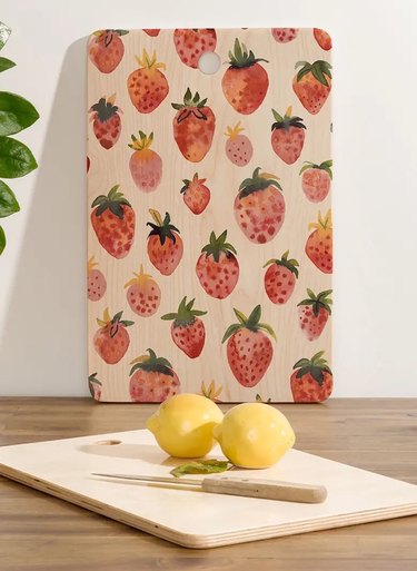 Deny Designs Strawberries Wood Cutting Board standing upright against a wall. Next to it is a plain wood cutting board with two lemons and a knife on it.