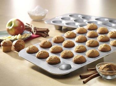 Mini muffins in a USA Pan mini-muffin pan, surrounded by ingredients including apples and cinnamon, with a standard-sized USA Pan muffin tin behind in blurred focus