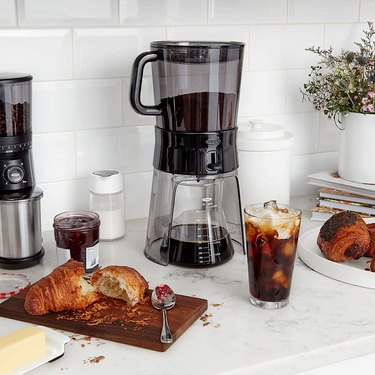 OXO cold brew coffee maker on a white marble countertop. The carafe looks like a glass beaker and the component that holds the coffee beans is plastic with a large handle. The beaker sits below the plastic component and there's a tap.
