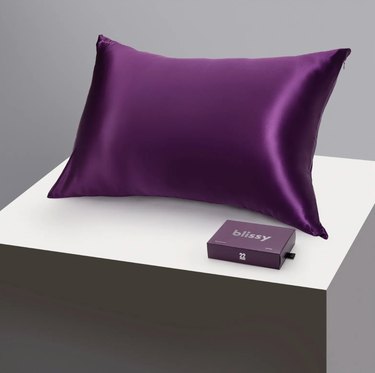 Blissy Mulberry Silk Pillowcase in purple pictured on a square stand next to the box it comes in.