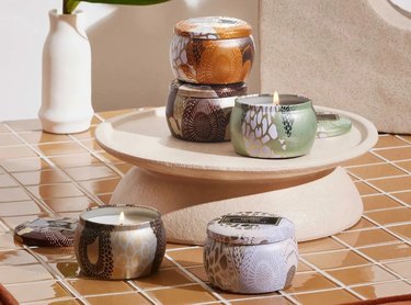VOLUSPA Japonica Set of 5 Mini Tin Candles sitting on and around a pedestal. The tins come in various colors and patterns.