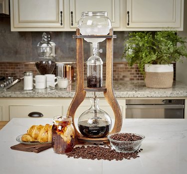 Nispira Cold Brew Drip Tower Coffee Maker pictured on a white countertop. It's about 18 inches tall and features a wooden stand and glass components.