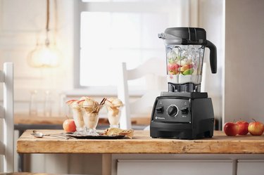 Vitamix 7500 Blender on a wooden countertop. There's ice and fruit in it and smoothie cups are placed next to it.