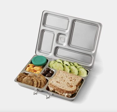 Open stainless steel lunch box with five compartments, full of lunch foods like a sandwich, crackers, and sliced cucumbers.