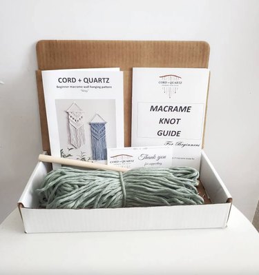 All the supplies in an open cardboard box. There's sage green robe, a wooden dowel, a knot guide, and an instruction booklet with photos of the finished product on the cover.