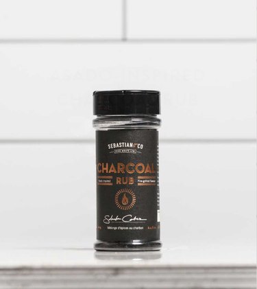 A plastic spice jar filled with charcoal rub sits on a countertop in front of white subway tiles.